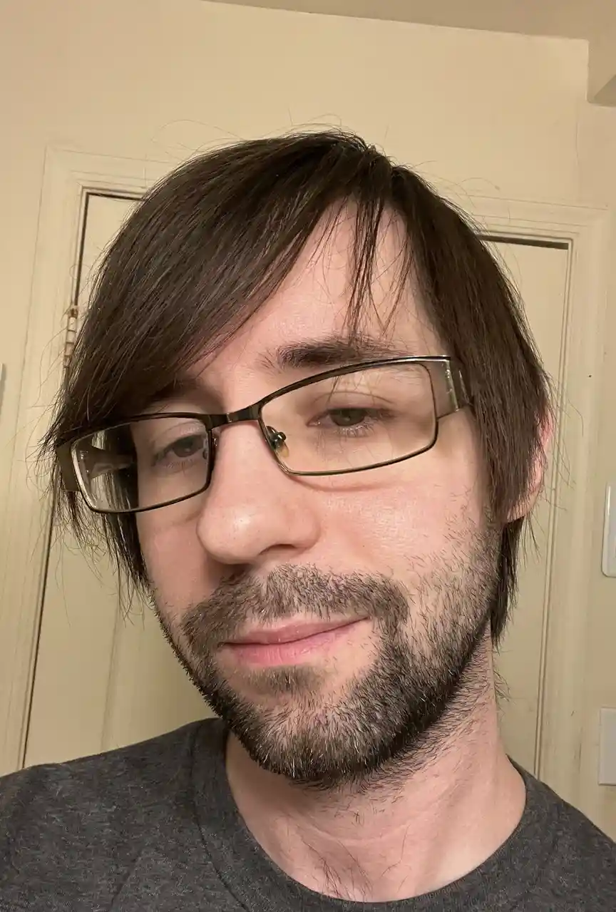 A close-up selfie of a man with dark hair, glasses, and a beard, wearing a gray t-shirt, looking at the camera with a slight…