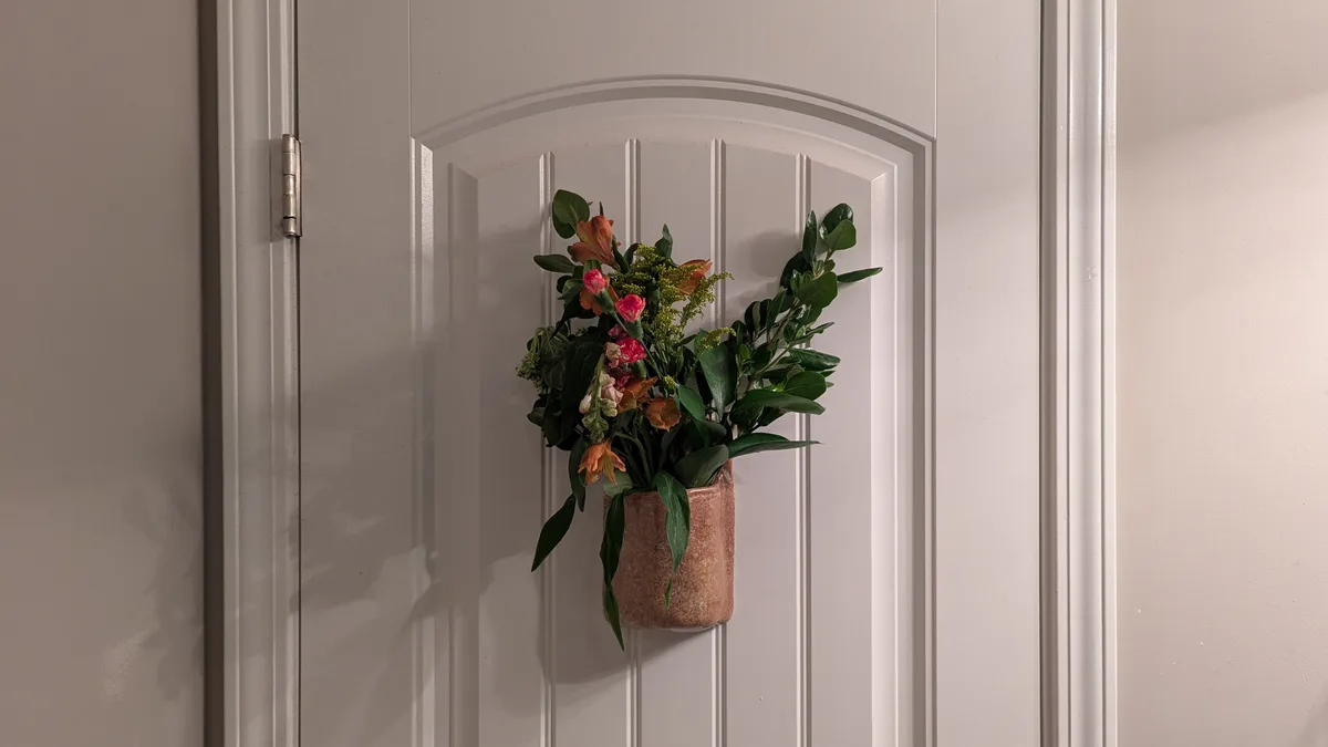 A decorative flower arrangement in a textured terracotta vase is mounted on a white paneled interior door, with the text…
