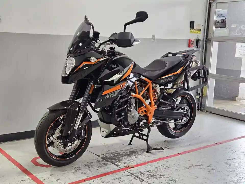 A black and orange KTM 990 Adventure motorcycle is parked indoors on a concrete floor marked with red lines. The bike…