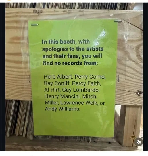 A bright green sign taped to a wooden record store shelf, listing famous artists whose records are intentionally absent from…