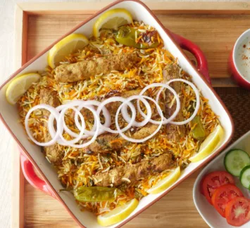 A close-up photo of a dish of biryani with meat, garnished with lemon slices and green chilies, topped with a decorative…
