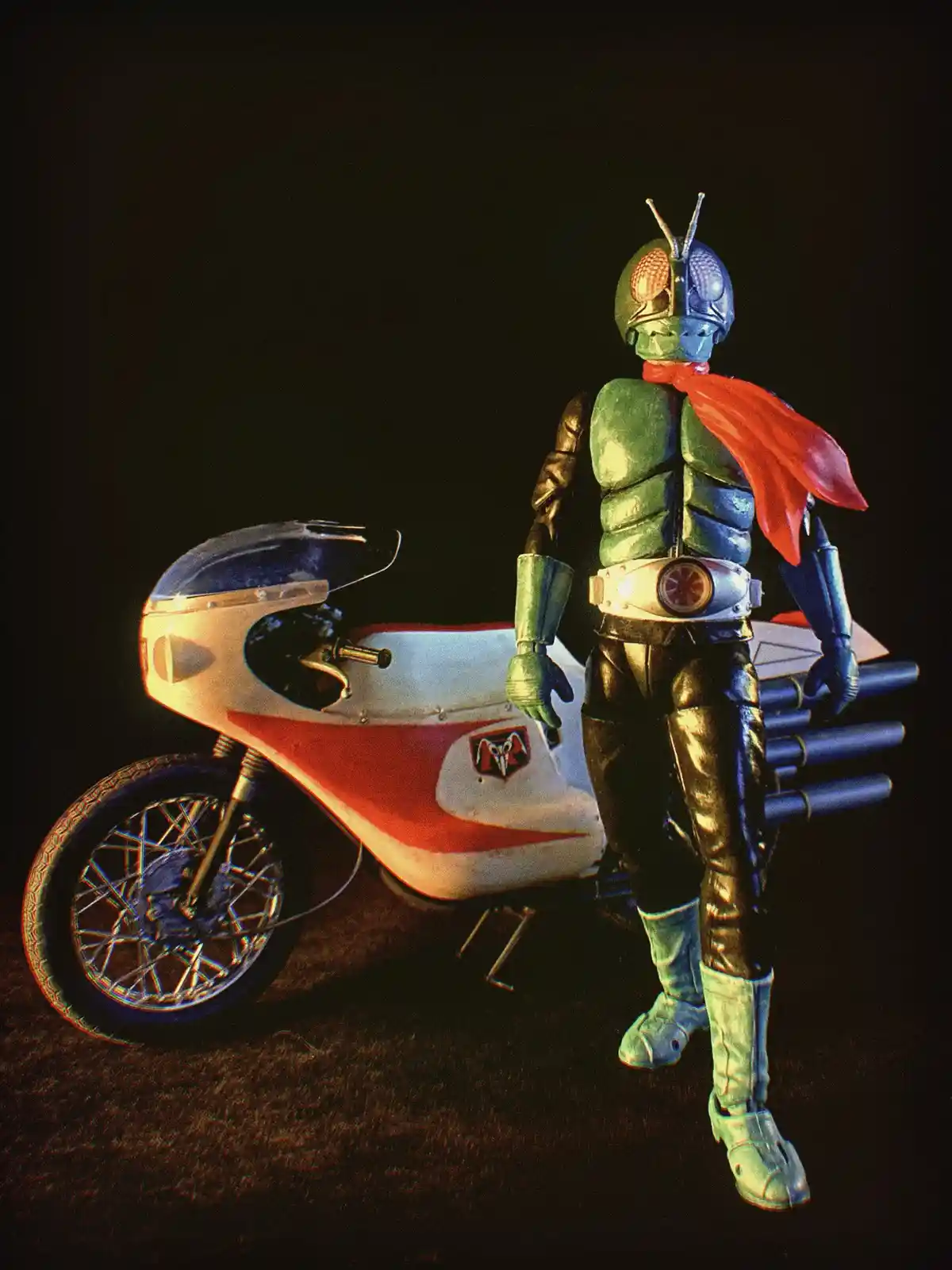 A figure in a green and blue armored costume with a red scarf stands beside a white and red motorcycle against a dark…