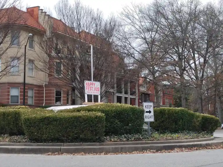 An outdoor photo shows a residential street with brick buildings and bare trees. In the foreground, a hedge surrounds a…