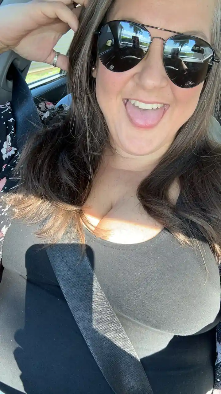 A woman wearing aviator sunglasses sits in a car, smiling and sticking her tongue out playfully while holding her hair. A…