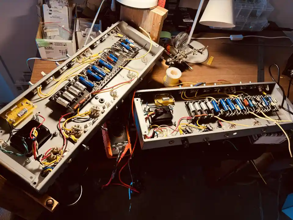 An overhead shot of a cluttered electronics workbench with two open metal enclosures revealing complex wiring, capacitors…