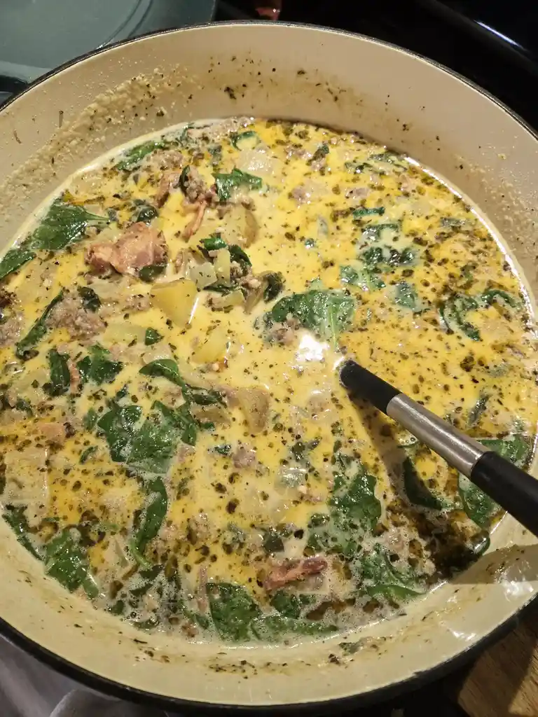 A close-up photo shows a creamy, yellowish dish in a light-colored pot, containing visible chunks of meat, green leafy…
