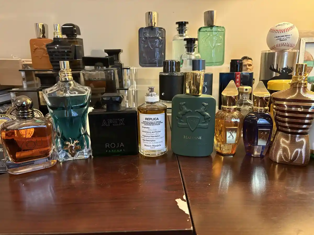 A wooden table displays a large collection of luxury perfume bottles with varied shapes, colors, and labels, including…