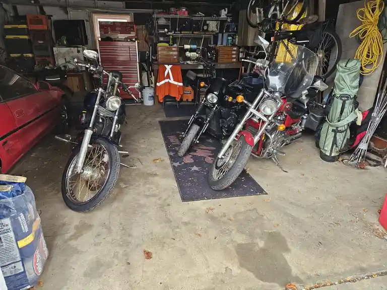A cluttered garage contains two motorcycles parked on a mat, a red car partially visible, and various tools, storage bins…
