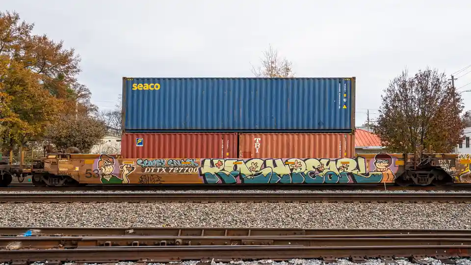 A freight train with two stacked shipping containers, a blue one on top and a red one below, sits on tracks with autumn…
