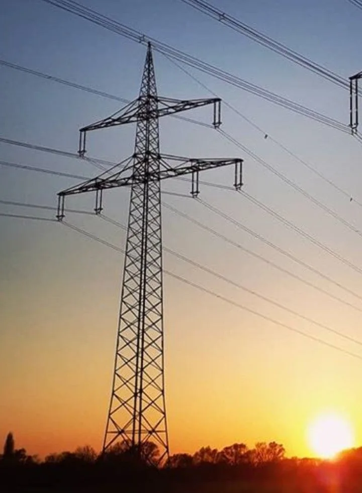 A tall, metal electricity pylon stands silhouetted against a sunset sky with orange and blue gradients, multiple power lines…