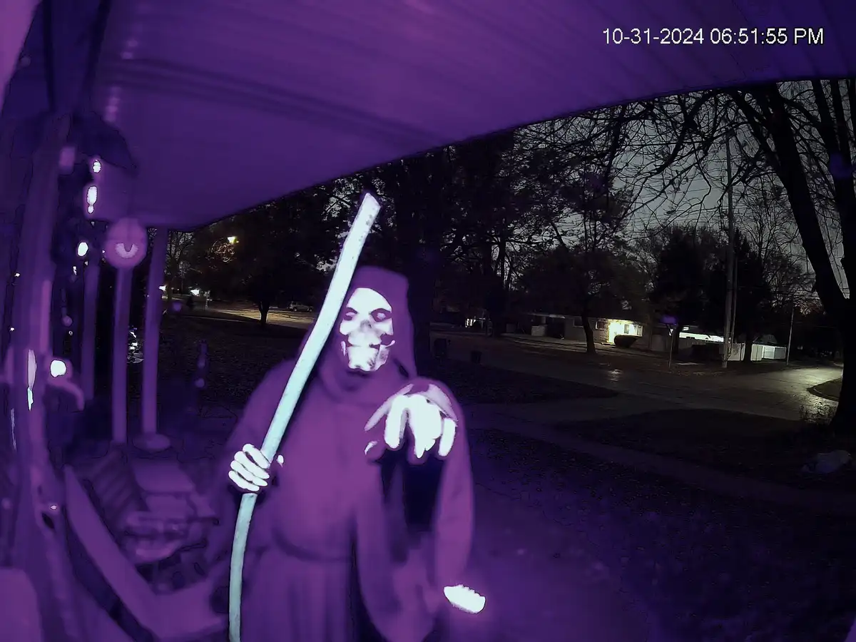 A security camera image shows a person in a grim reaper costume with a skull mask and a long staff, standing outside at…