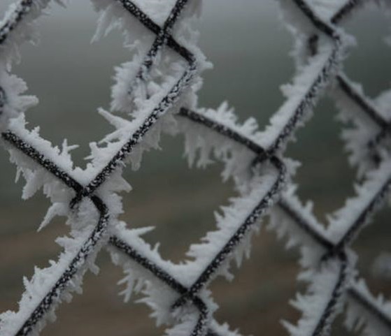 A close-up photo of a chain-link fence covered in intricate, frosty ice crystals, with a blurry, muted background. Overlaid…