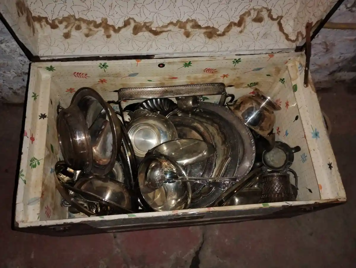 An open, worn wooden box with floral-patterned lining contains a jumble of tarnished silverware including plates, bowls…