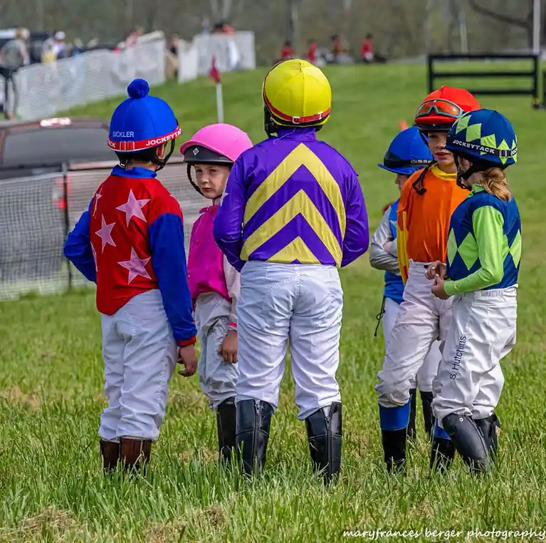 A group of young children dressed in colorful jockey silks and helmets stand on a grassy field, facing away from the camera…