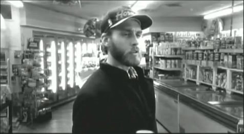 A black-and-white image of a man with a beard and a baseball cap standing in a convenience store aisle, looking slightly…