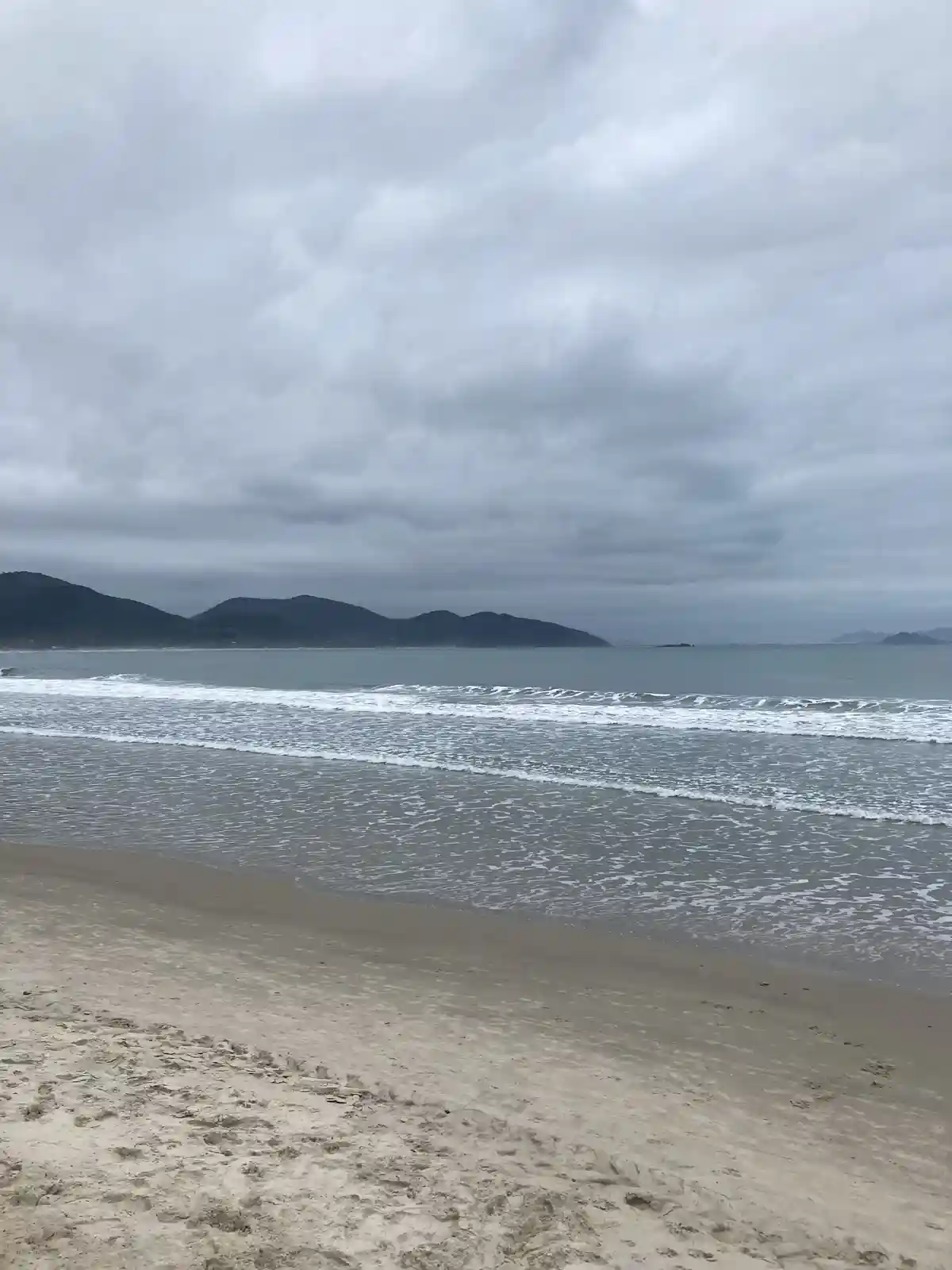 A wide shot of a beach under an overcast sky, with gentle waves rolling onto the sand and distant hills on the horizon. The…