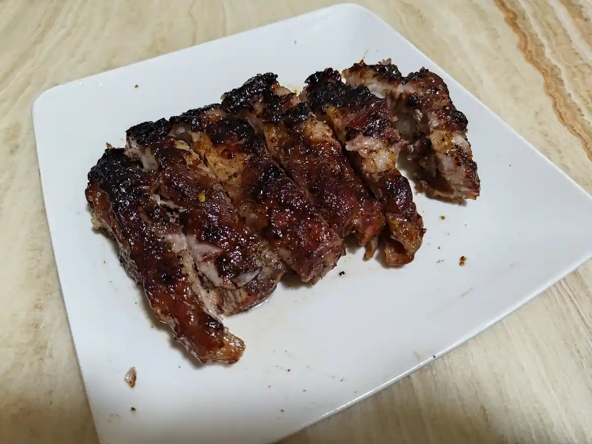 A white square plate holds sliced, charred meat with a glossy glaze, resting on a light wood-grain surface. The overlaid…