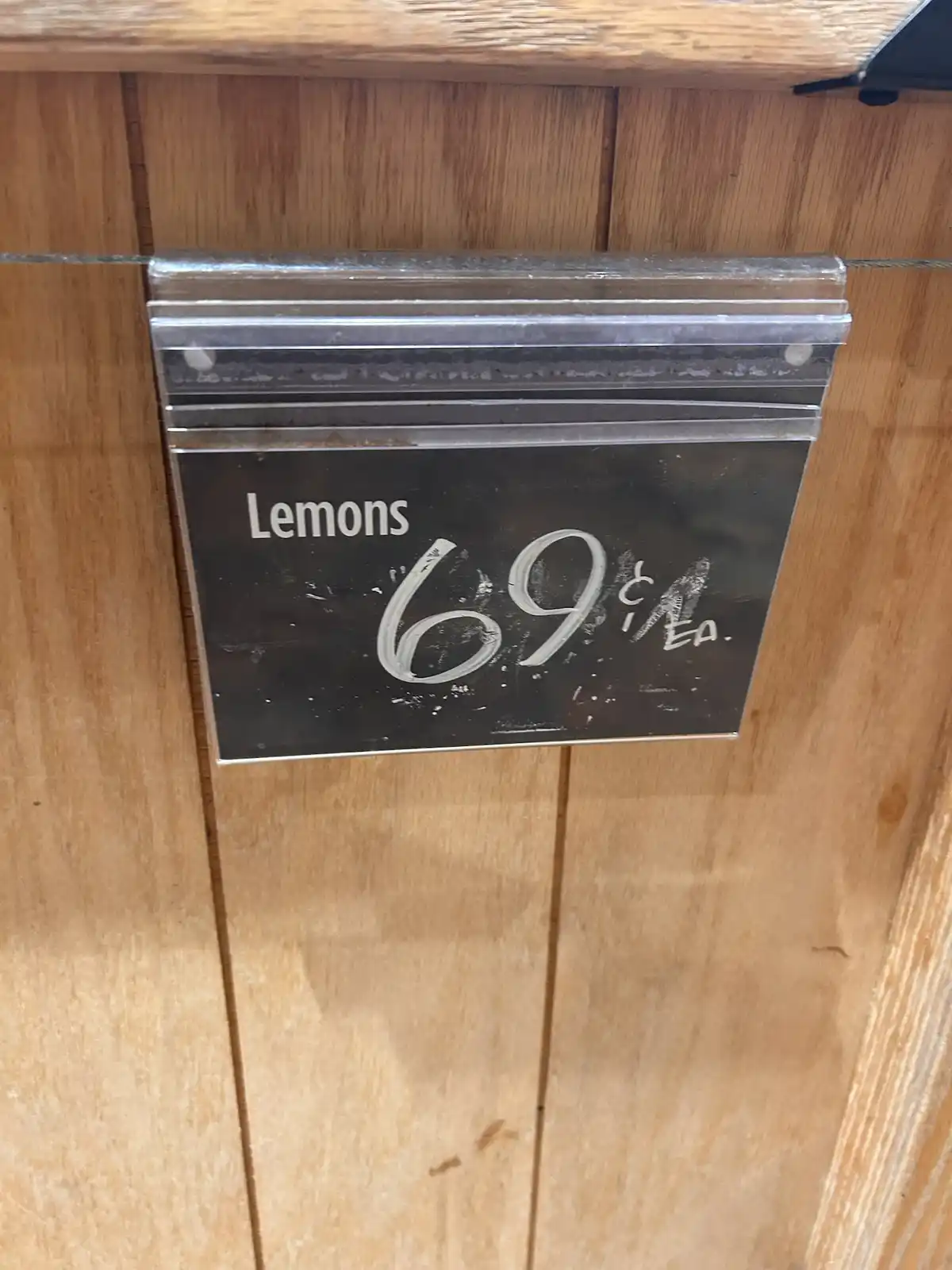 A close-up of a black price tag in a wooden display case shows the word 'Lemons' printed in white, with '69¢ EA.' written in…