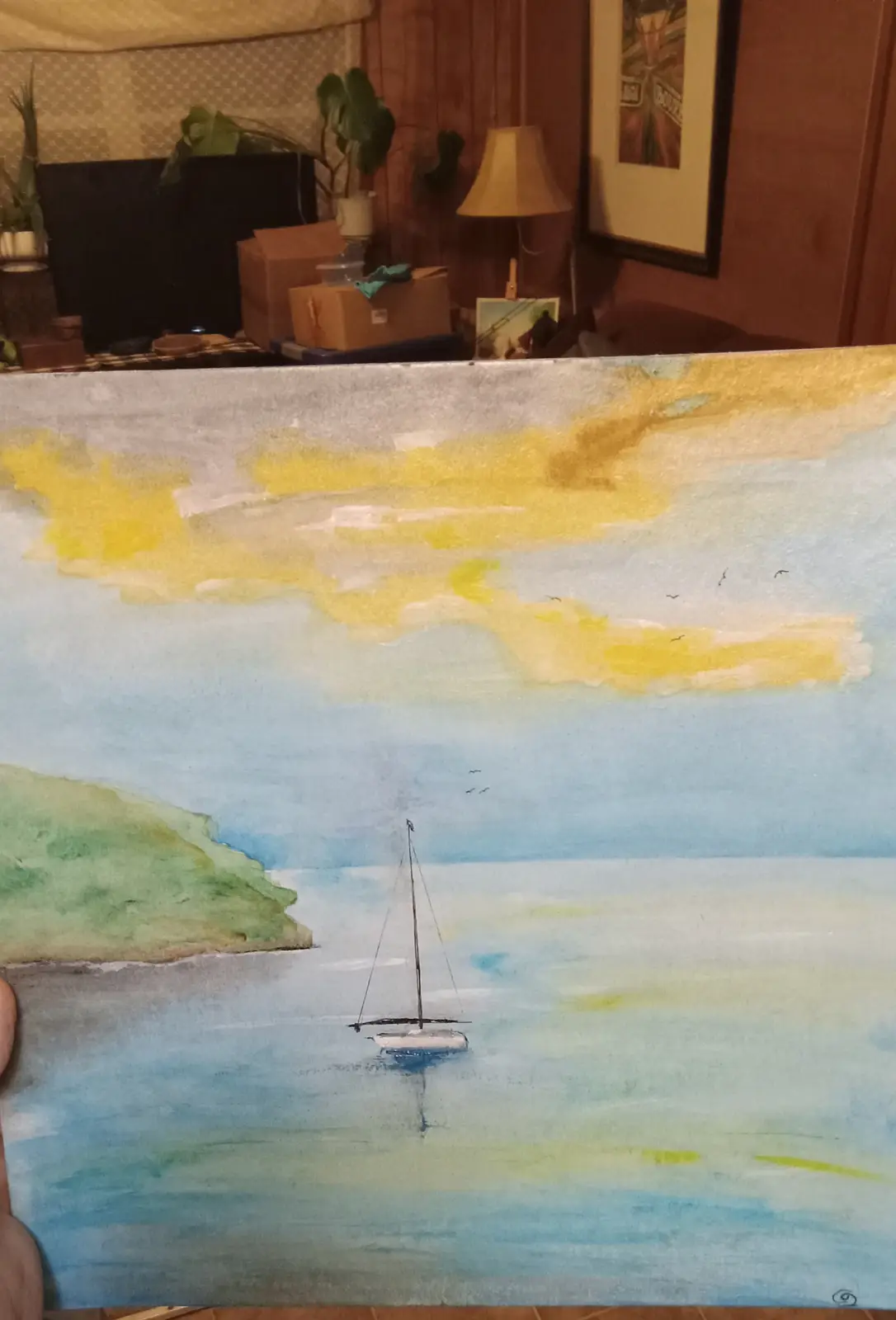 A person holds up a watercolor painting depicting a sailboat on calm blue water near a green landmass under a sky with…