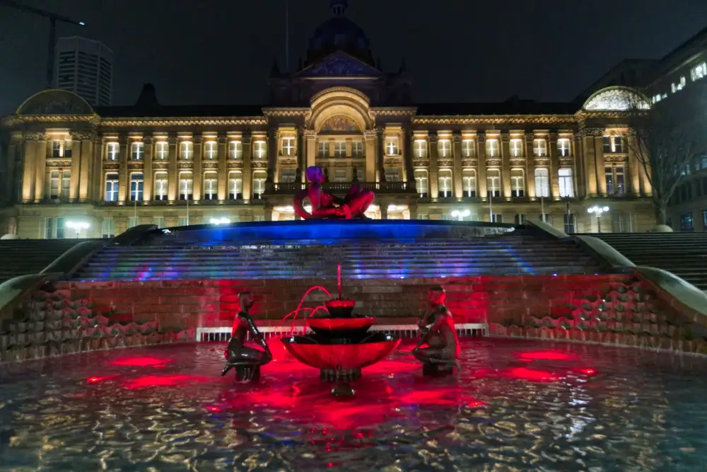 A nighttime photo of a grand, illuminated building with a large fountain in front. The fountain is lit with red and blue…