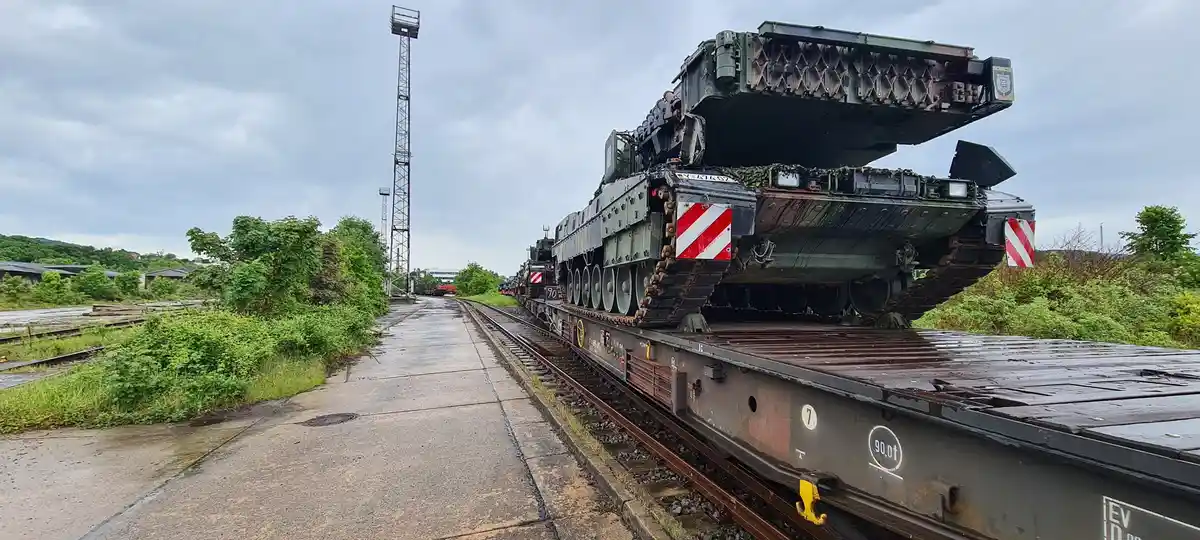A large military tank is mounted on a flatbed train car, traveling along railway tracks under an overcast sky. The tank is…