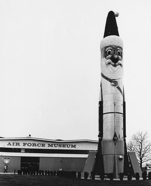 A black-and-white photo shows a large rocket painted to resemble Santa Claus, complete with a hat, beard, and smiling face…