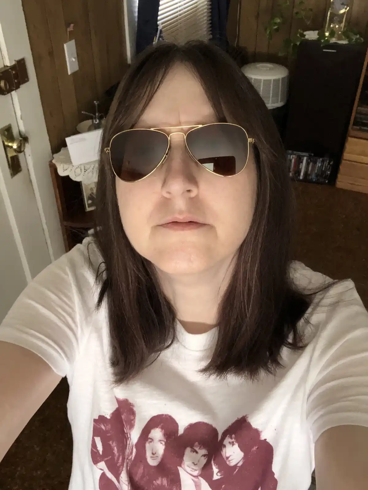 A person with shoulder-length brown hair wearing gold-framed aviator sunglasses and a white t-shirt with a red band graphic…