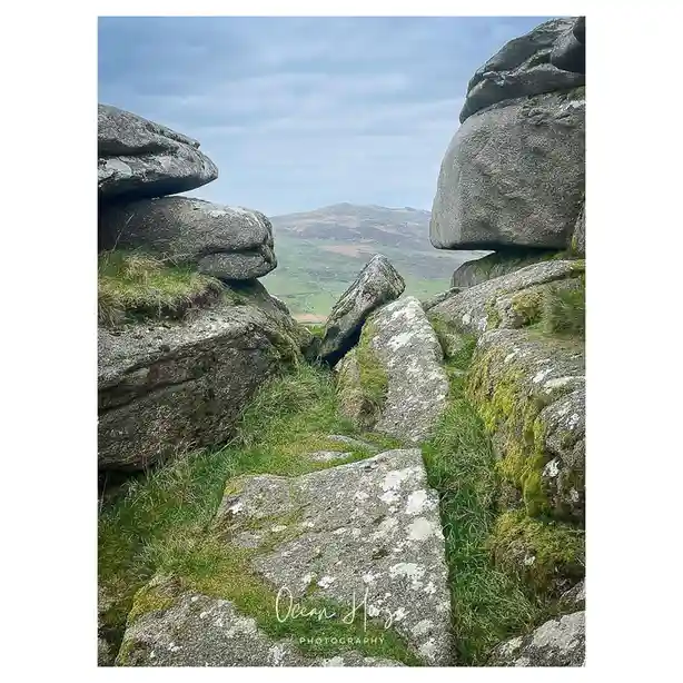A scenic photo of a narrow rocky path between large, moss-covered boulders, leading toward a distant green hillside under a…