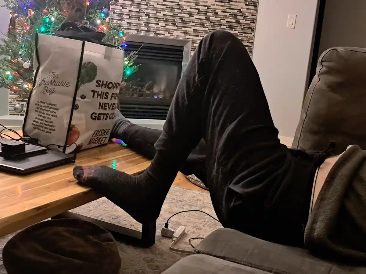 A person lounging on a couch with their feet up on a wooden coffee table, next to a laptop and a large reusable shopping bag…