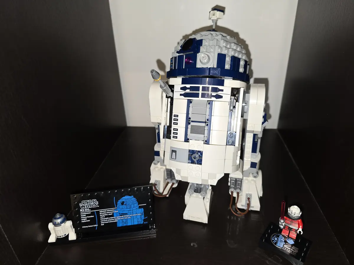 A large, detailed LEGO model of the Star Wars droid R2-D2 stands on a dark shelf, flanked by a small R2-D2 minifigure and a…