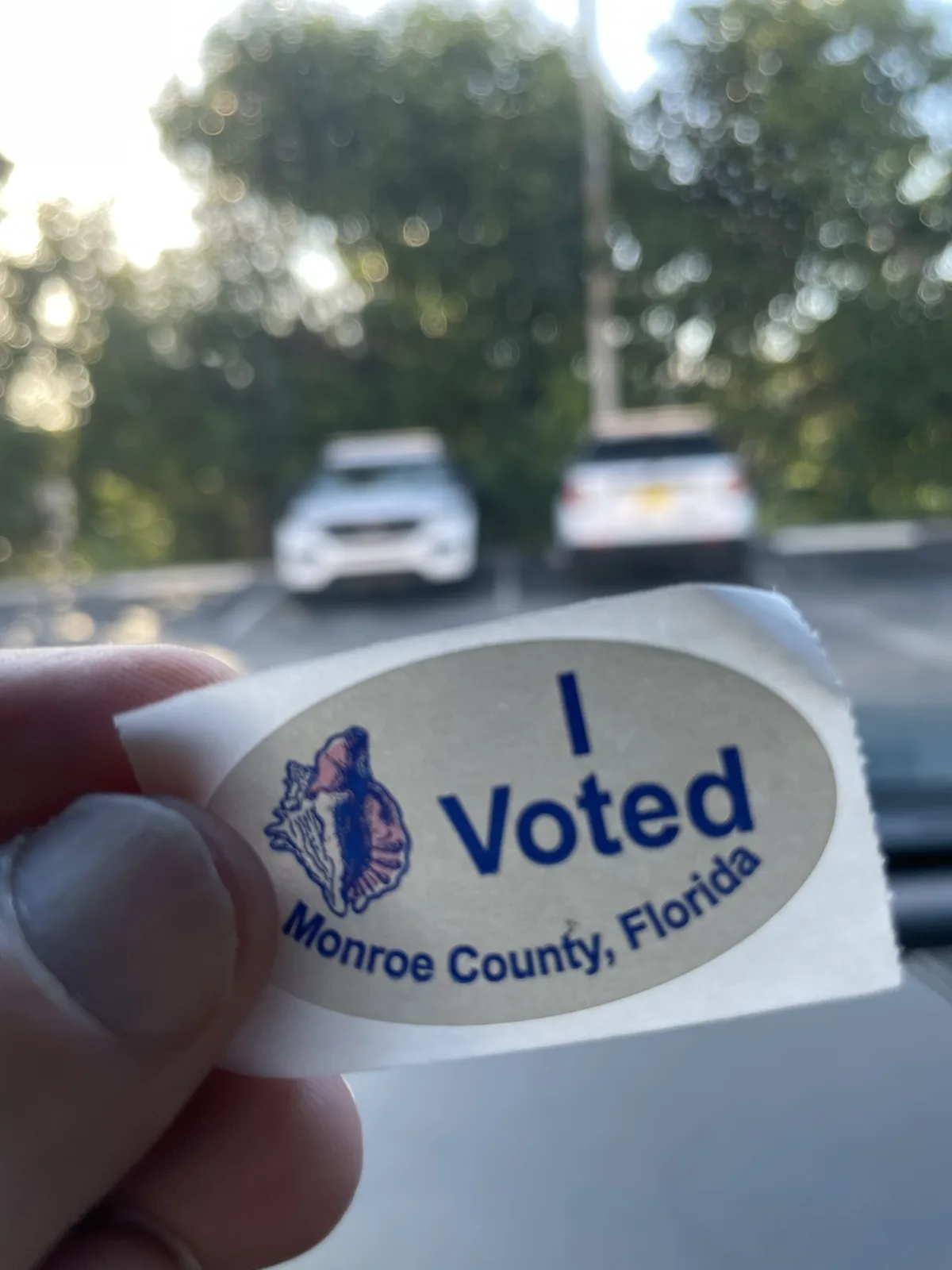 A hand holds up a beige oval sticker that reads 'I Voted Monroe County, Florida' with a seashell logo, taken from inside a…