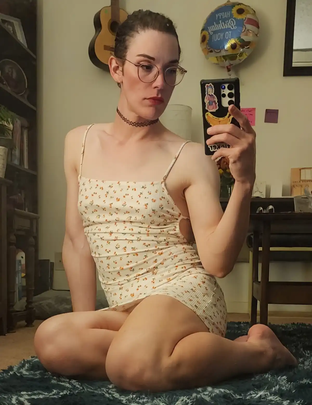 A person with glasses and a floral dress sits cross-legged on a rug, taking a mirror selfie with a phone that has banana and…