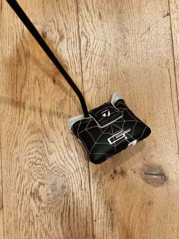 A black golf putter head with a quilted cover and white stitching lies on a light brown wooden floor. The putter has a black…