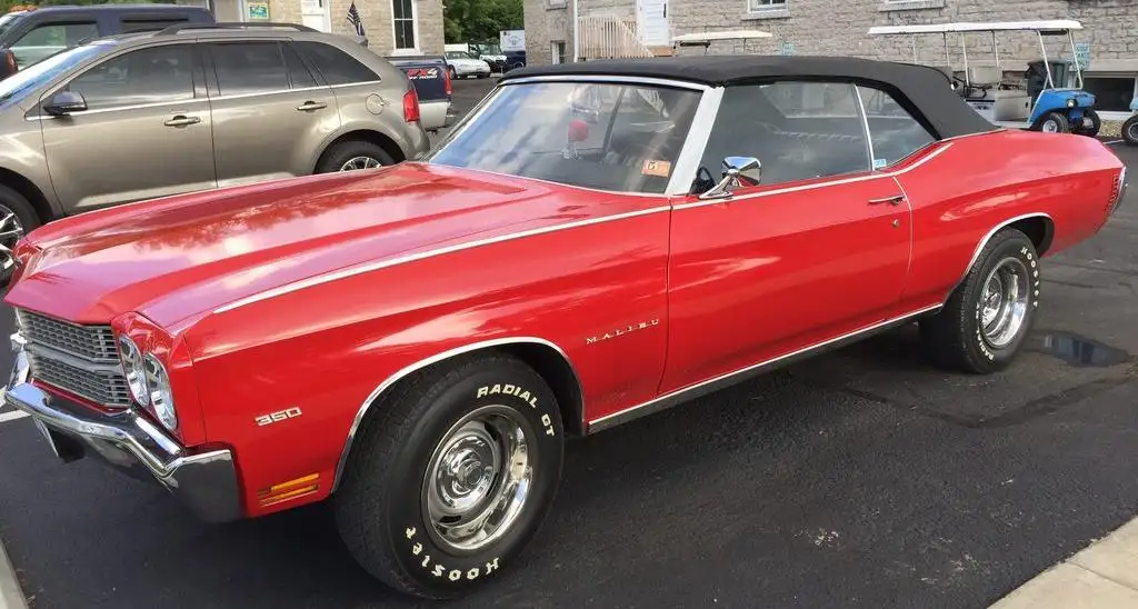 A shiny red 1960s Chevrolet Malibu convertible with a black soft top parked in an asphalt lot, surrounded by modern…