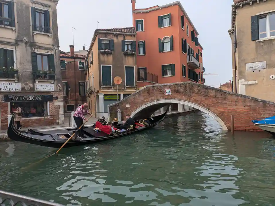 A gondolier in a red-and-white striped shirt rows a black gondola with ornate gold trim through a Venetian canal, passing…