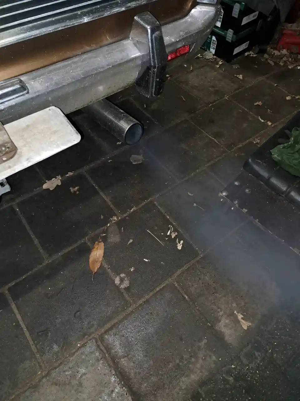 A close-up photo shows smoke billowing from the exhaust pipe of a brown truck parked on a dark tiled floor scattered with…