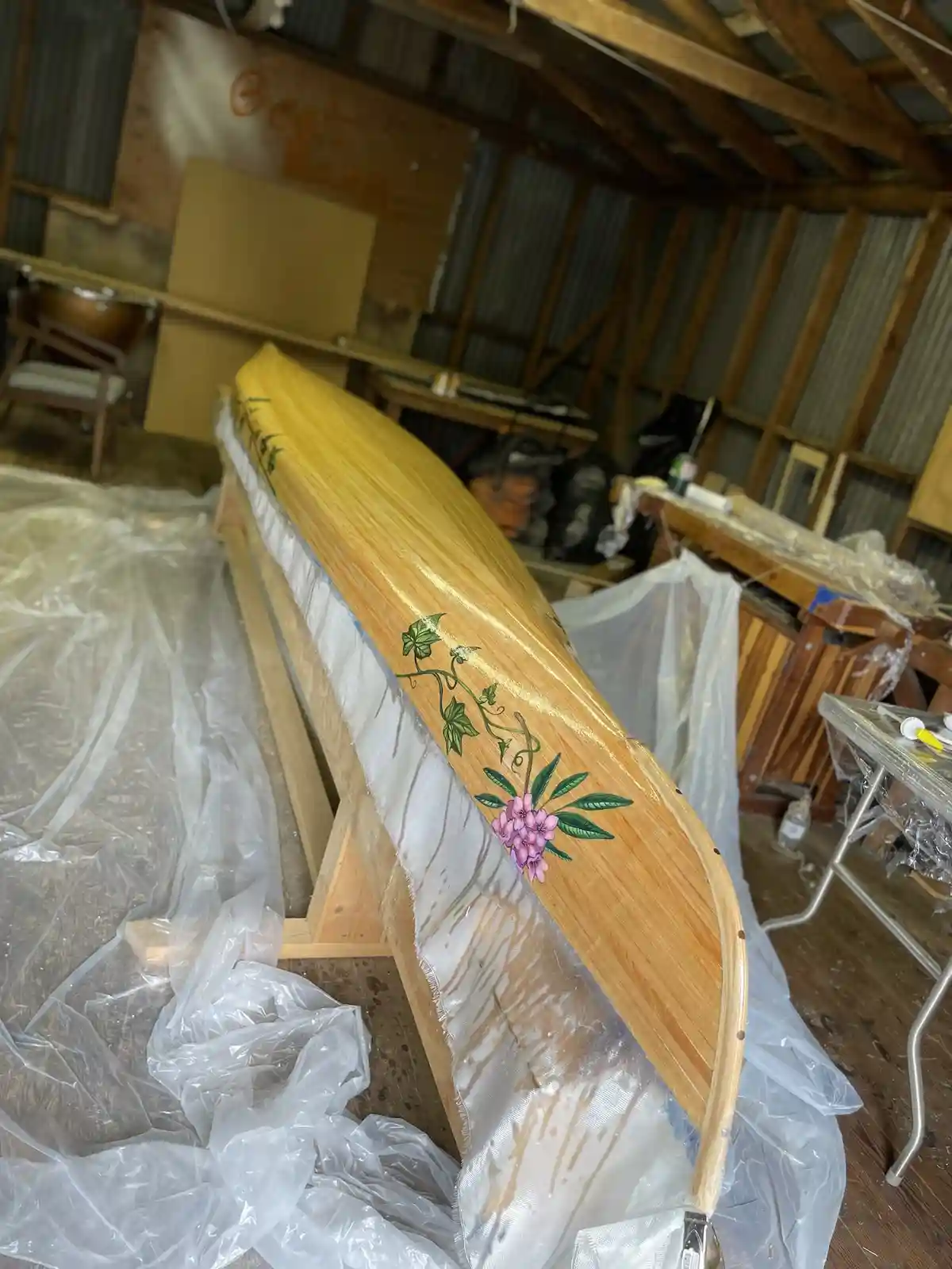 A wooden canoe rests on a stand in a workshop, partially covered in plastic sheeting. It features a hand-painted floral…