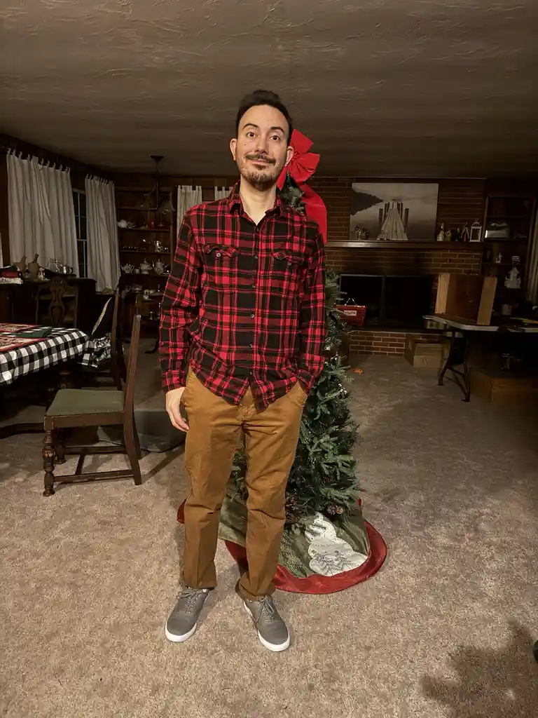 A man in a red and black plaid shirt and brown pants stands in a living room next to a small, decorated Christmas tree. The…
