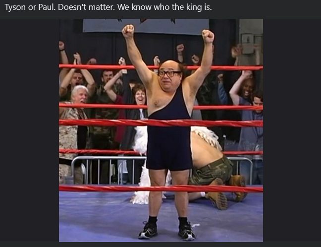 A man in a dark singlet and glasses stands triumphantly in a wrestling ring with arms raised, while another man in…