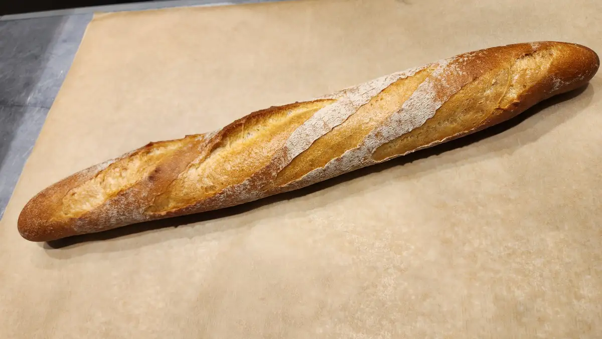 A freshly baked baguette with a golden-brown crust and flour dusting lies diagonally on parchment paper atop a stainless…