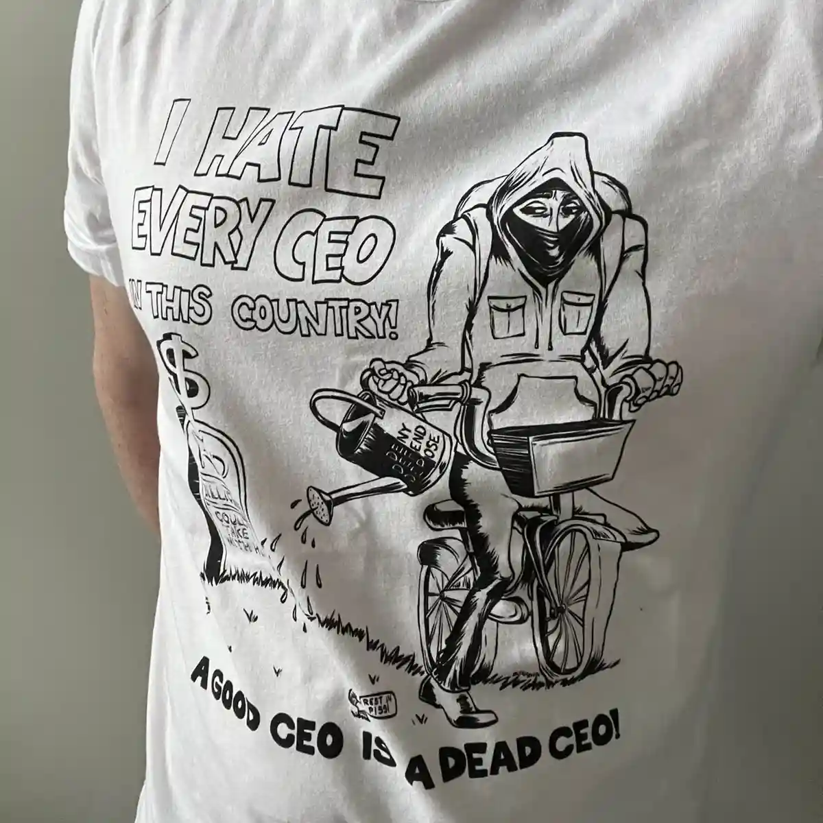 A white t-shirt features a black-and-white cartoon of a hooded figure on a bicycle watering a grave marked with a dollar…