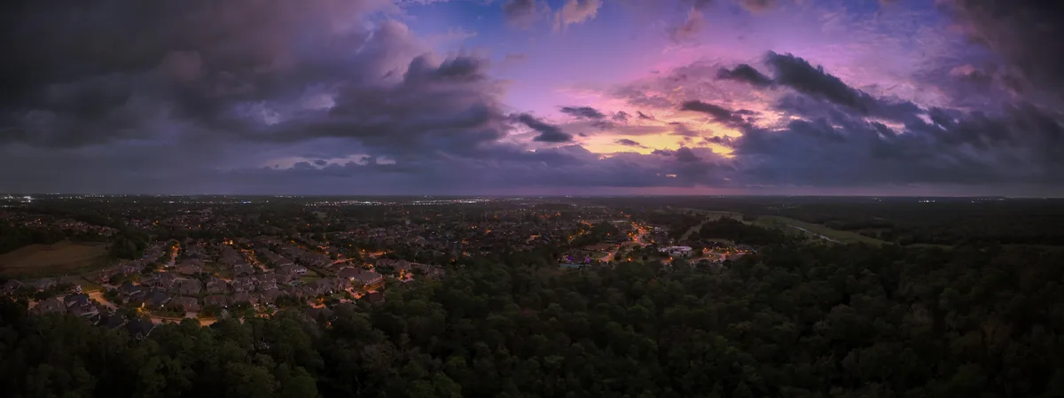An aerial panoramic photo captures a sprawling suburban neighborhood at dusk, nestled between dense forests, under a…