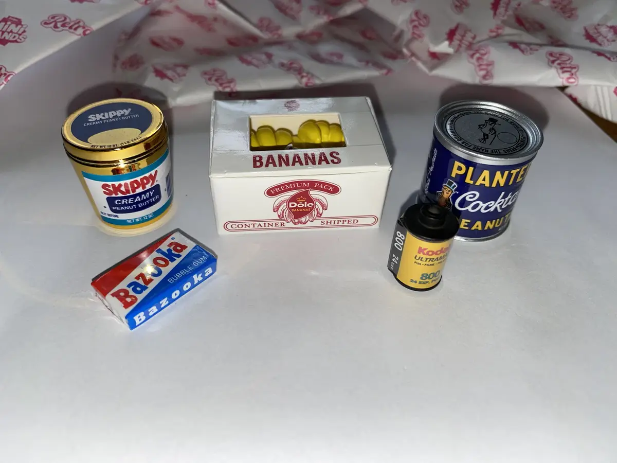 A still life of five miniature, vintage-style items arranged on a white surface: a Skippy peanut butter jar, a Dole bananas…