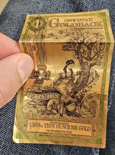 A hand holds a gold-colored, rectangular Utah Goldback token, labeled as 'Voluntary, Local Currency' and stating it contains…