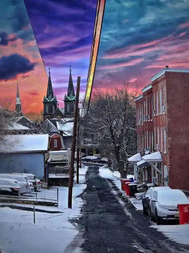 A snowy street scene with brick buildings, parked cars, and snow-covered trees, framed by a dramatic, colorful sunset sky…