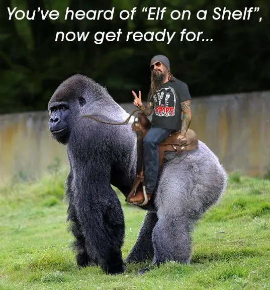 A man with a beard, sunglasses, and a black t-shirt sits atop a large silverback gorilla in a grassy area, making a peace…