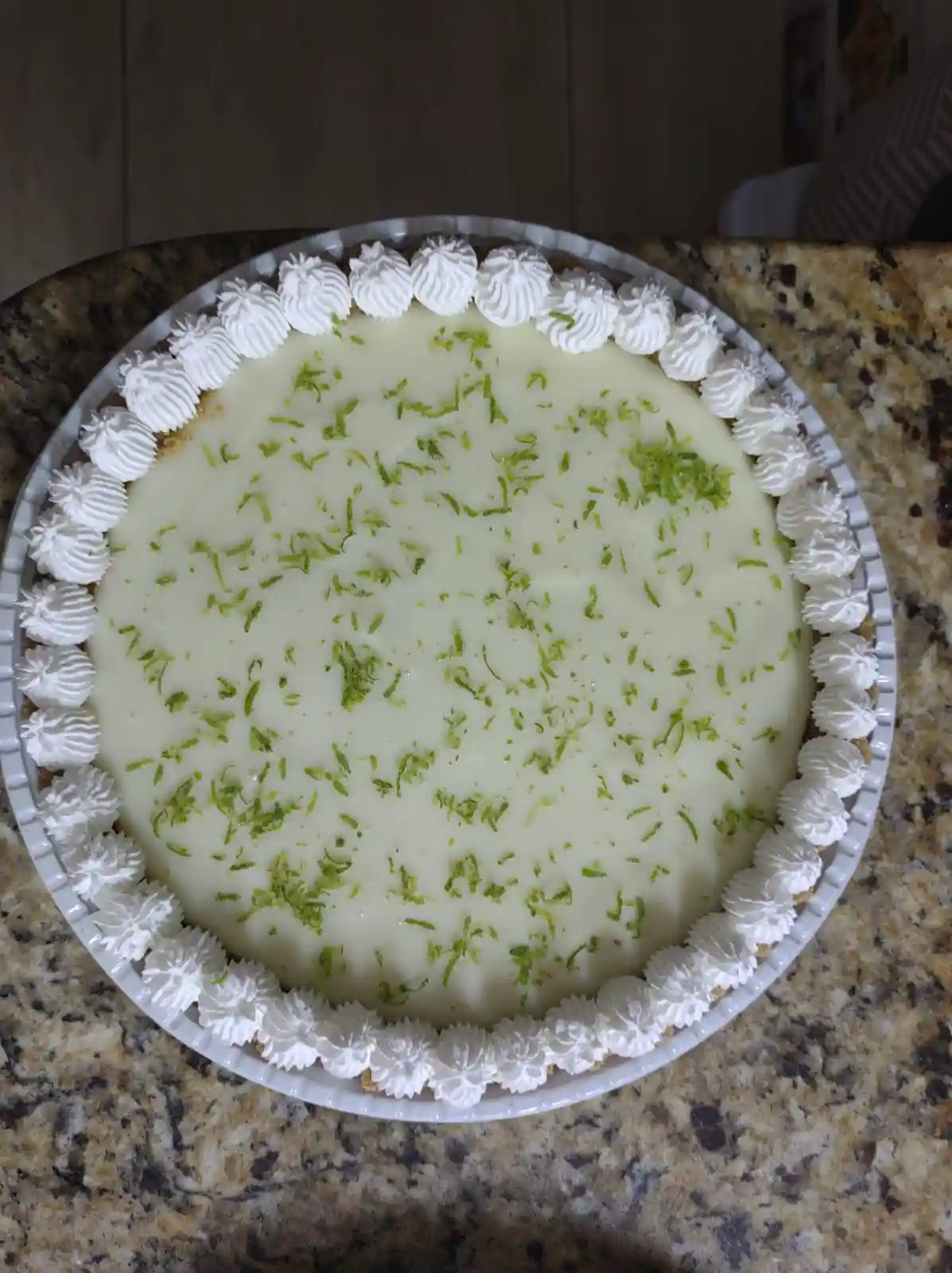 A top-down photo of a round, creamy lime pie in a white fluted dish, garnished with a border of piped whipped cream and…