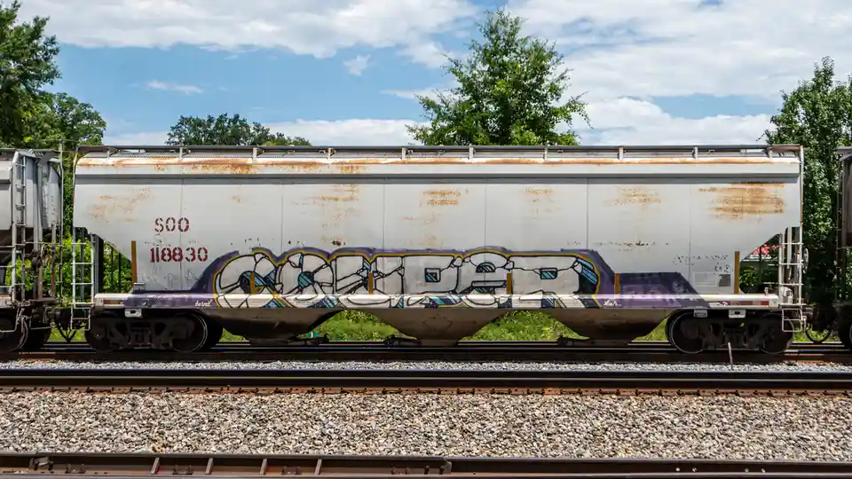 A white, rusty railcar with graffiti reading 'CAGED' in stylized lettering sits on train tracks under a partly cloudy sky…