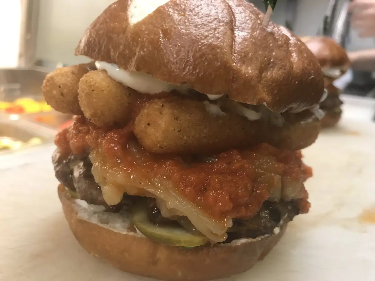 A close-up photo of an extremely tall, messy burger with a beef patty, melted cheese, pickles, a thick red sauce, and a…