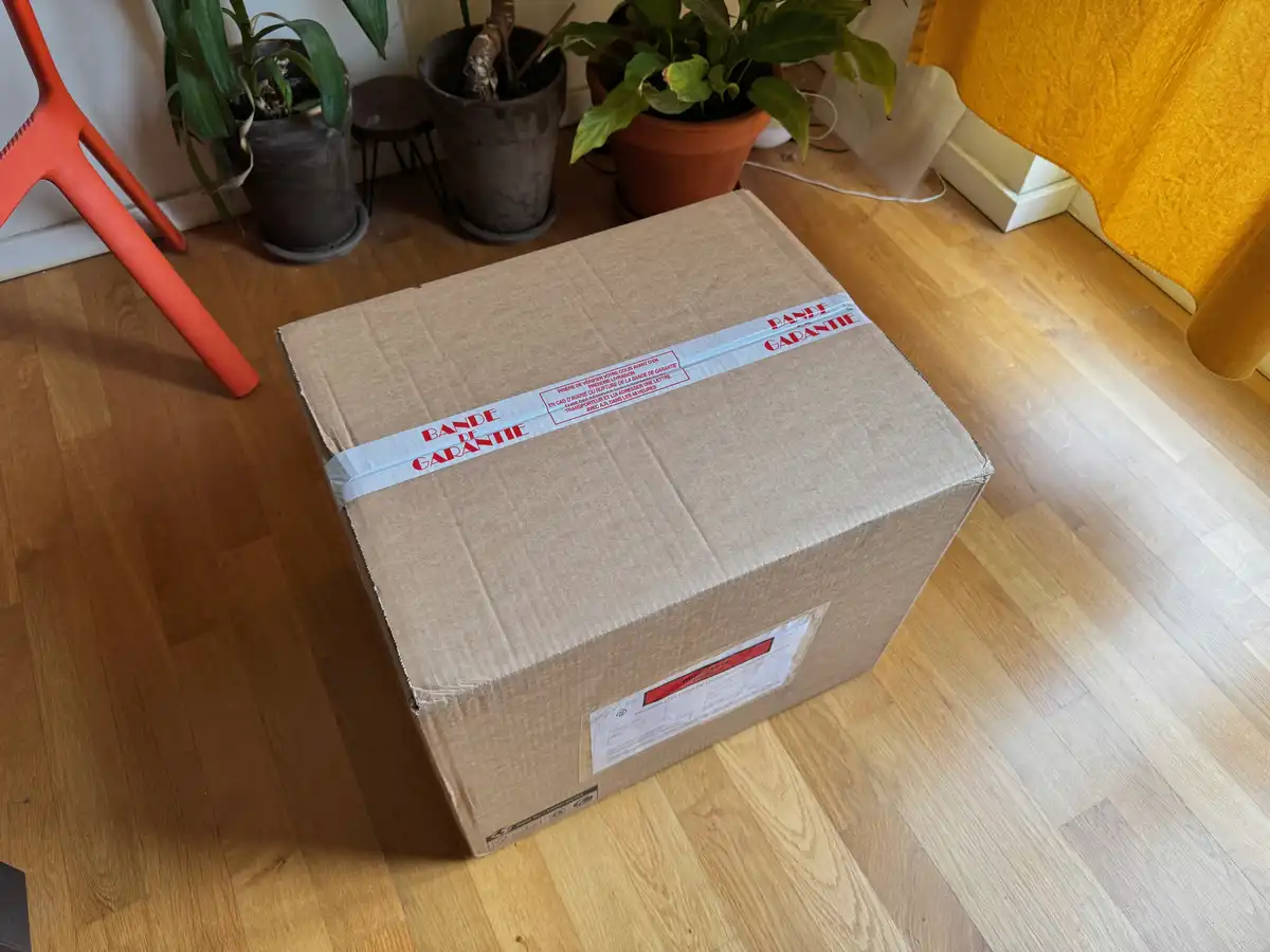 A large cardboard shipping box sits on a wooden floor, secured with a white tape that reads 'BANDE DE GARANTIE' in red…